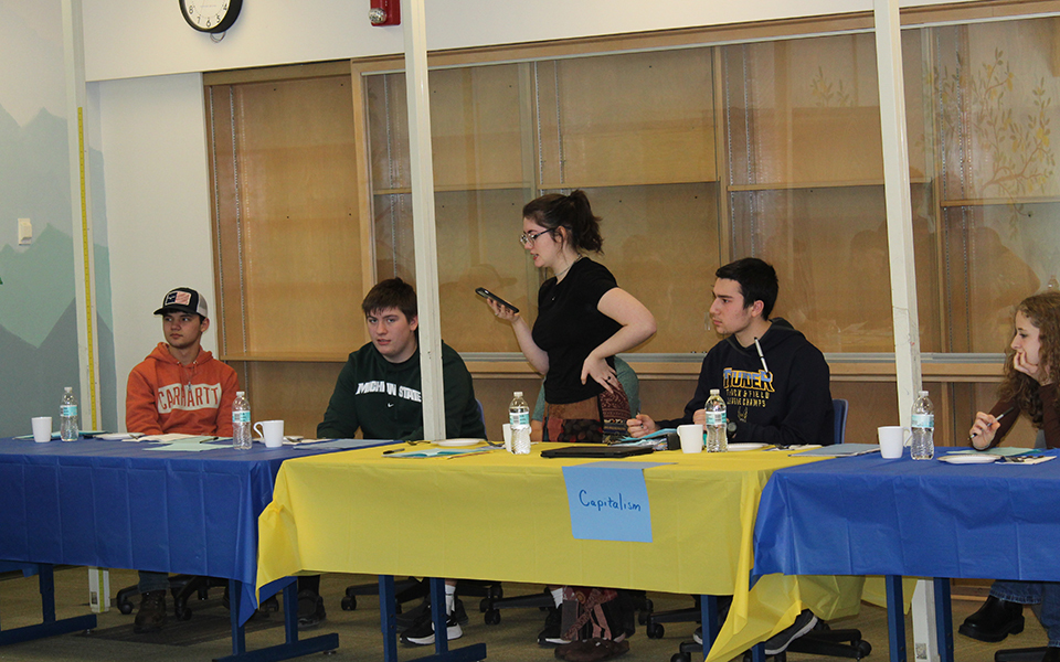 students seated behind tables draped with plastic tablecloths. One standing, hand on hip