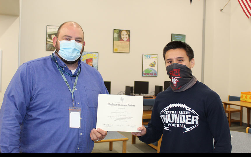 2 people indoors, masks, holding certificate