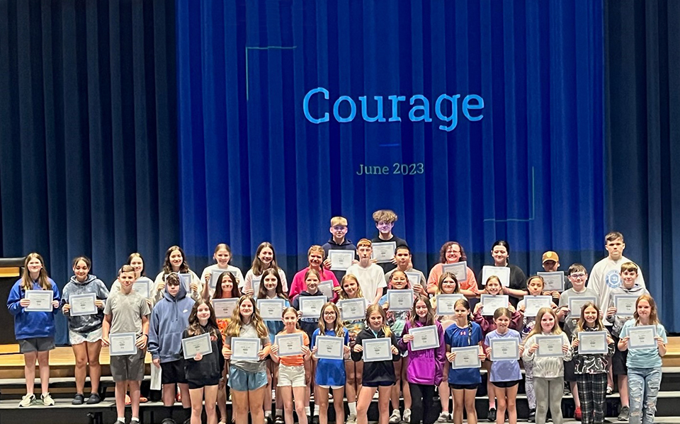 Group of students standing on auditorium riser, holding certificates, word courage projected behinds