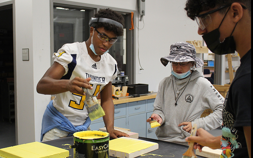 three students in tech class painting stool tops yellow