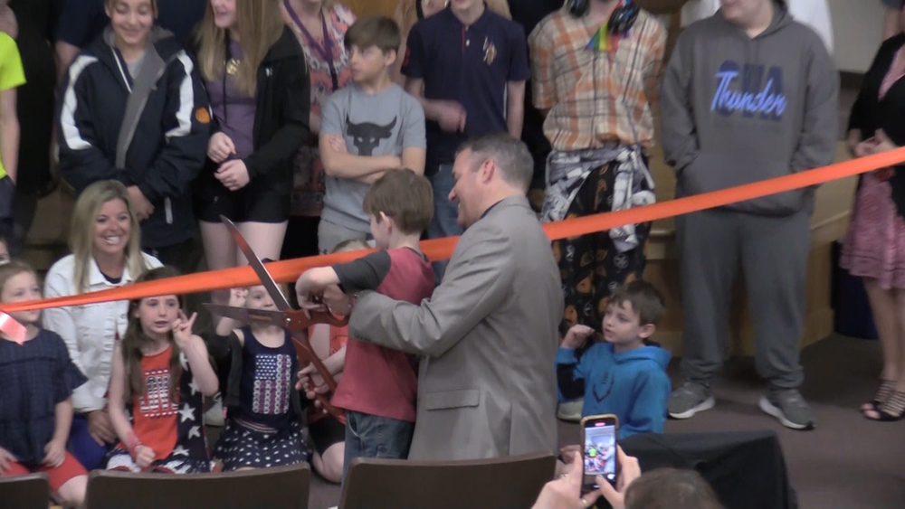 man and small boy cut a ribbon with a giant pair of scissors while crowd of students watch