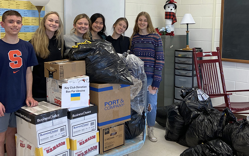 classroom, six students standing, cart filled with boxes, floor covered with large garbage bags
