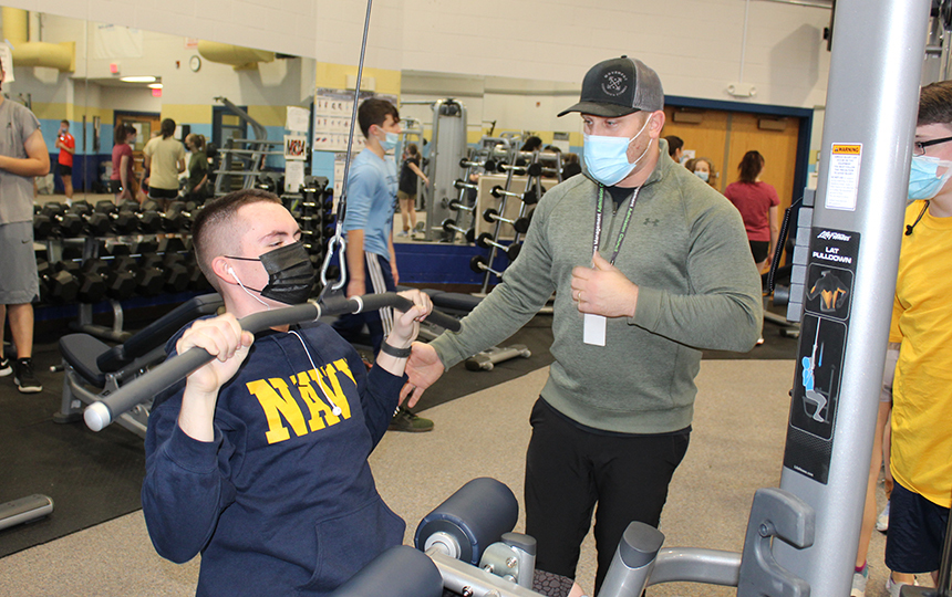 Fitness center, student seated on weight machine, man standing beside instructing