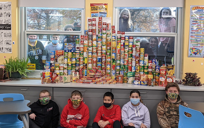 Stacks of canned goods on table in classroom, students seated in front. students peering in window behind