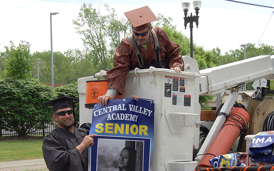2 men in cap and gown, one in boom lift, hanging banners