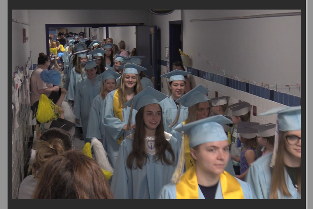 Students in cap and gown walking down hallway lined with young students