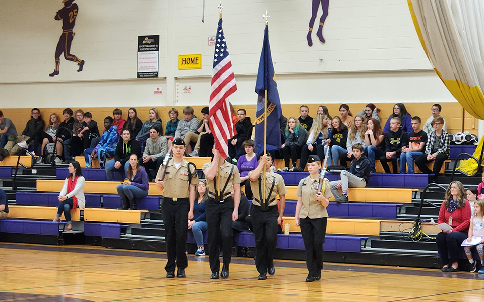 gym, bleacher with students, four cadets, uniforms, flags, drill rifles