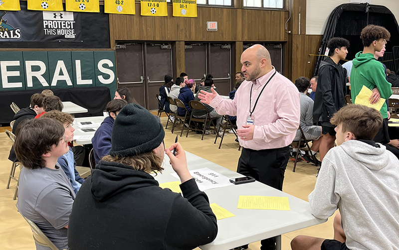 gymnasium, man standing speaking, students seated at tables.