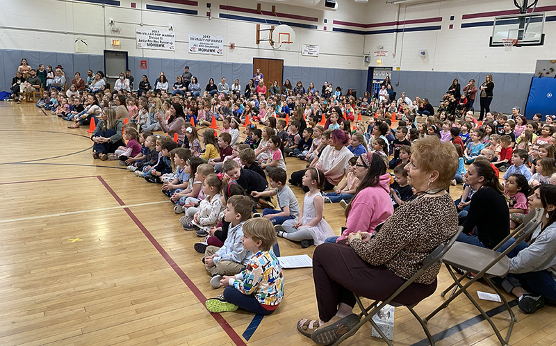 hundreds of students seated on the floor, teachers in chairs, gym