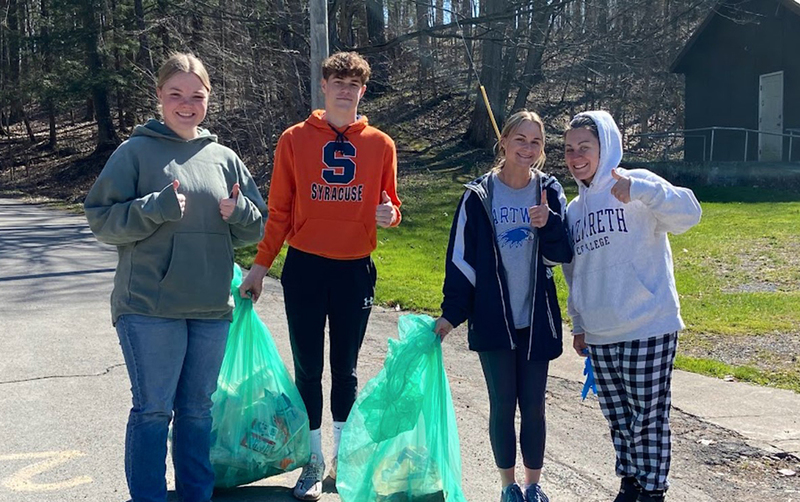 4 students outside with trash bags, sidewalk, grass, trees