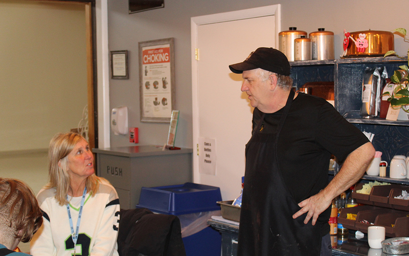 man in apron speaking to woman seated at table, cupboard, sign, garbage can