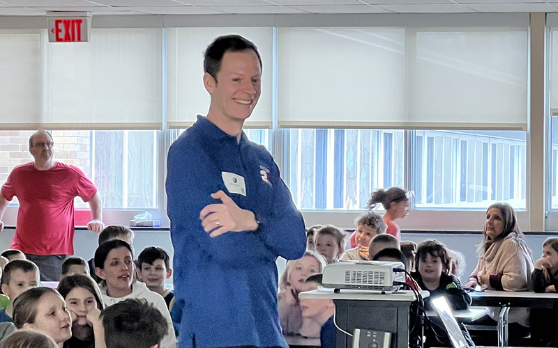 man smiling, arms crossed, students seated at tables, bright windows