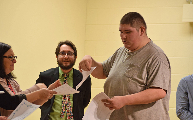 tall student taking papers from woman, man watching and smiling