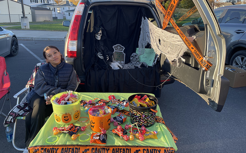 girl seated at table filled with candy, car with open trunk, outdoors
