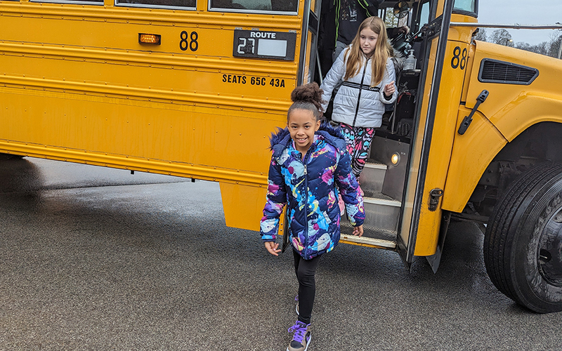 2 girls smiling, disembarking school bus, outdoors, coats