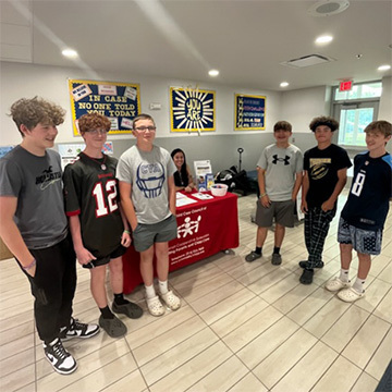 6 boys standing, woman seated at table, lobby, windows, posters