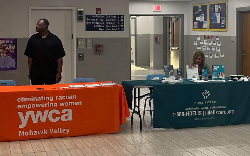 man standing at table draped with YMCA banner, woman seated at table draped with fideliscare.or banner, doorway, hall